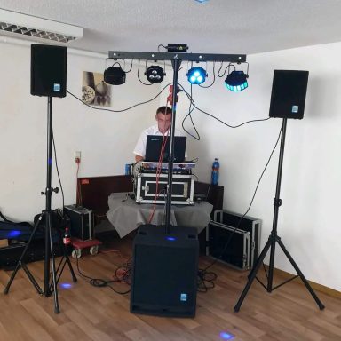 Ein DJ steht hinter einem Mischpult mit Lautsprechern und Lichteffekten in einem Raum.
